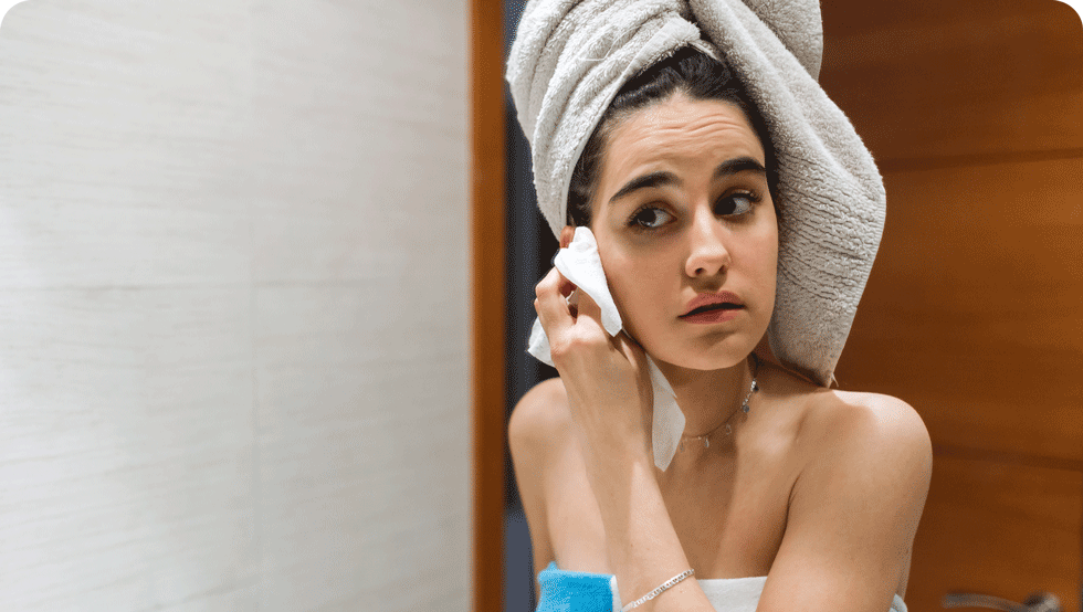 A young woman with a bath towel on her head cleansing her face after getting out of the shower